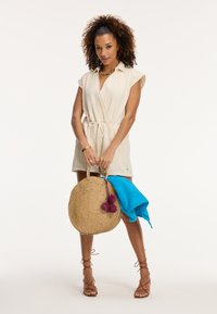 Kremowa sukienka w stylu wrap z krótkim rękawem, wykonaną z teksturowanej tkaniny, z wiązaniem w pasie. Brązowa pleciona torba ze słomy z pomponami, na której leży turkusowy ręcznik.
