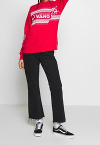 Sudadera roja con un patrón geométrico blanco y el logo "VANS", combinada con unos vaqueros acampanados negros y zapatos negros con suelas blancas.