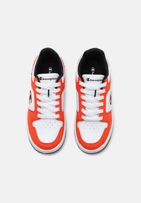 Baskets blanches et orange avec des accents noirs. Tige en cuir lisse, boîte à orteils perforée et lacets blancs. Logo Champion sur le côté.