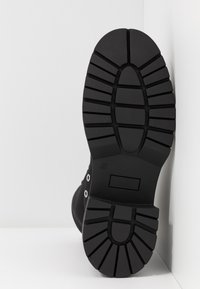 Semelle de botte noire montrant une surface en caoutchouc texturée avec des crampons profonds pour l'adhérence. Présente des découpes circulaires et ovales près de la zone des orteils. Taille 37 imprimée.