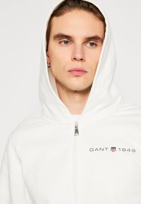 Felpa bianca con zip realizzata in tessuto morbido, caratterizzata da un logo con "GANT 1949" e uno scudo in nero e rosso. Il cappuccio aggiunge calore.