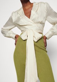 Kremowa plisowana bluzka z wciągnięciem na przodzie, związywana z przodu, zestawiona z oliwkowo-zielonymi wysokimi spodniami o prążkowanej fakturze, z długimi rękawami i kołnierzykiem.