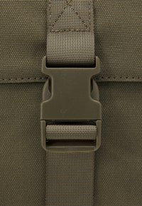 Sangle en tissu vert olive avec une boucle en plastique. Surface texturée avec des détails de couture et motif de renforcement triangulaire sur la sangle.