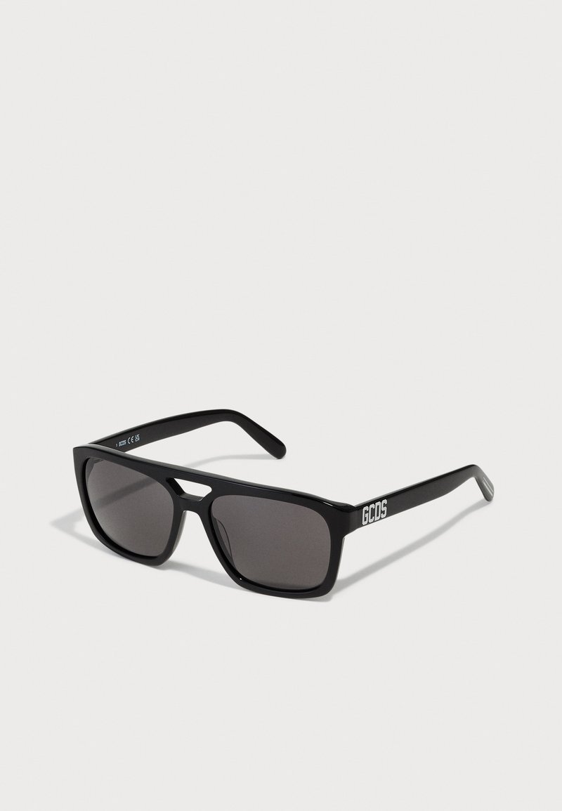 Gafas de sol negras con montura cuadrada y lentes oscuros, con un logo de GCDS en las patillas. Fabricadas en plástico con una textura suave.