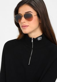 Pulover negru cu fermoar, cu textură canelată, având un guler înalt și logo-ul brodat "OBEY". Ochelari de soare aurii, tip aviator, cu lentile gradient.
