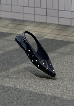 Fekete hegyes orrú szandál ezüst szegecsekkel, aszfalt fölött lebegve, csempézett fal háttérrel.