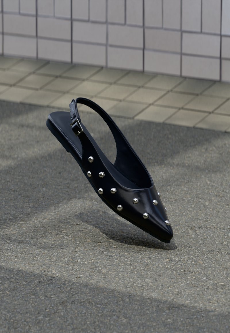 Μαύρο μυτερό παπούτσι slingback με ασημένια τρουκ, να αιωρείται πάνω από άσφαλτο κοντά σε πλακιδωτό τοίχο.