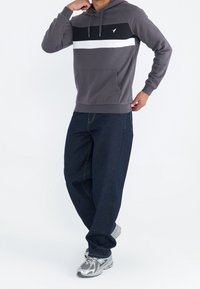 Hombre con sudadera gris con franjas en blanco y negro, jeans oscuros y zapatillas plateadas, de pie con una mano cerca de la cara.