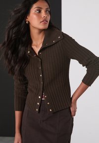 Cardigan marrone a coste con ampio colletto, caratterizzato da bottoni dorati, maniche lunghe e design aderente, abbinato a una gonna scura.