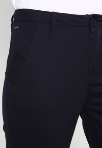 Navy blå bomullsblandade byxor med smal passform, med en framknapstängning och snedställda sidofickor. Slät textur och minimal detaljering.