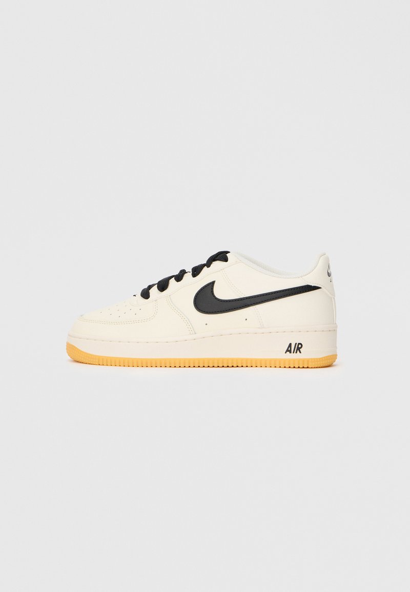 Tênis Nike Air Force 1 em couro creme com detalhes e cadarços pretos, apresentando uma sola de borracha texturizada e a marca "AIR" no calcanhar.
