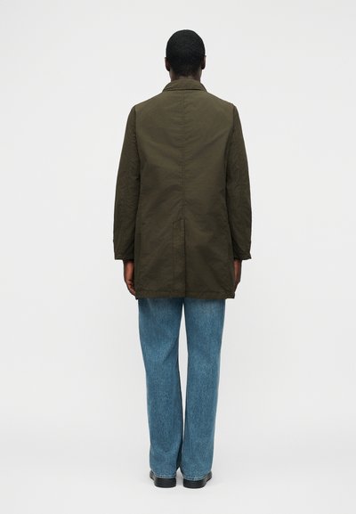 Άνθρωπος που στέκεται γυρισμένος με την πλάτη, φορώντας μακρύ μπουφάν ελιάς, μπλε τζιν και μαύρα παπούτσια, πάνω σε απλό φωτεινό φόντο.