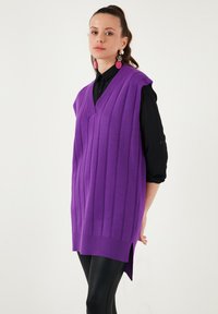 Purple ribbed sleeveless sweater with a v-neck, side slits, and a relaxed fit. Worn over a black, button-up shirt and paired with leggings.