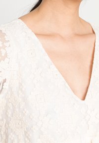 Blusa bianca ricamata con scollo a V, motivi geometrici e un tessuto trasparente e testurizzato con dettagli intricati.