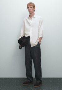 Weißes, knöpfbares Hemd, dunkel gestreifte Hose, braune Wildleder-Loafers, hält eine braune Tasche. Glattes Material, lockere Passform, modernes Design.
