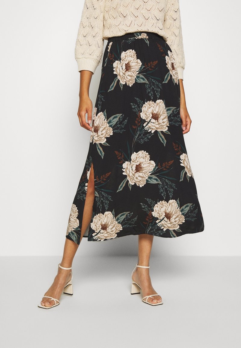 Femme portant une jupe midi noire avec de grands imprimés floraux beiges et une fente latérale, associée à des sandales blanches à talons à brides et un pull beige.
