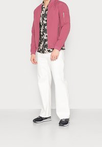 Giacca bomber leggera rosa con zip frontale e tasche laterali, sopra una camicia nera a fiori. Abbinata a pantaloni bianchi e sneakers nere.