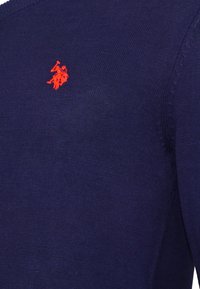 Tummansininen neuloskangas, jossa on pieni punainen brodeerattu poloilijatunnus vaatteiden vasemmassa yläkulmassa.