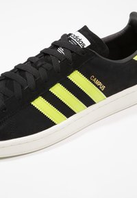 Svarta mockaskor med tre djärva neongula ränder, en vit gummisula och guldtexten "CAMPUS" på sidan. Svarta snören och tunga.