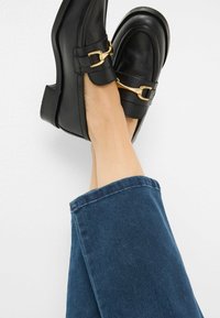 Mocassins en cuir noir avec un détail de boucle dorée, dotés d'un petit talon carré, associés à un jean en denim bleu foncé.