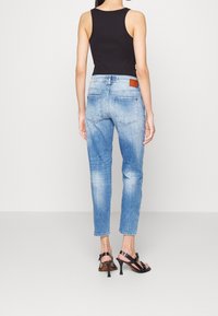 Light blue denim jeans with a tapered leg, featuring back pockets and a brown leather patch at the waist. Worn with a black tank top.