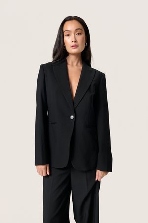 Schwarze Blazer mit Reverskragen, einreihiger Knopfschluss und zwei Fronttaschen. Taillierte Passform aus strukturiertem Stoff, kombiniert mit schwarzen Hosen.