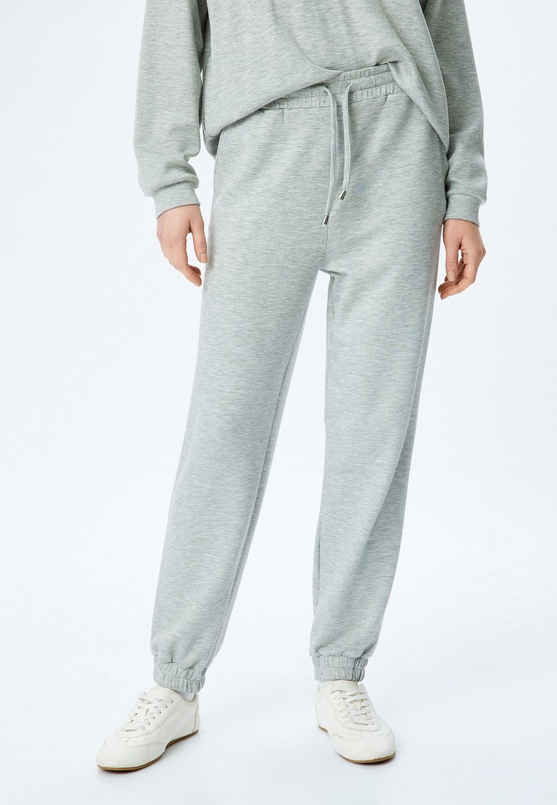Grijze sweatpants van zacht materiaal met een elastische tailleband en manchetten. Voorzien van een trekkoord en zijzakken. Gepaard met witte sneakers.