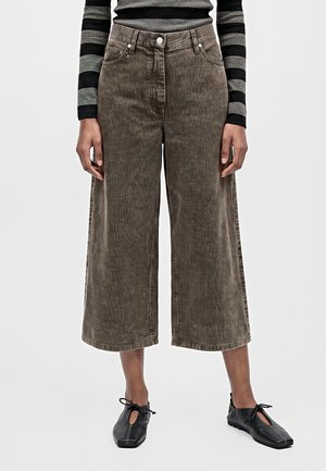 Un mannequin porte un jean marron court à jambes larges, un haut rayé noir et gris et des chaussures noires à lacets, sur un fond uni.