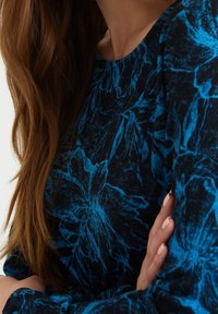 Top de tela negra con un diseño floral azul, mangas largas y cuello redondo. Textura suave con un estilo ajustado.