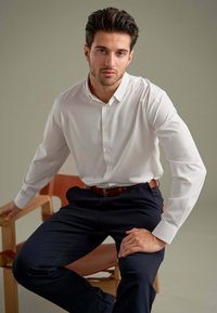 Hvid skjorte med knapper, lavet af bomuld, med lange ærmer, parret med mørkeblå bukser. Modellen sidder på en træstol med læderdetaljer.
