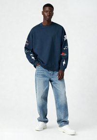 Sweatshirt bleu marine avec des designs brodés sur les manches, featuring des symboles comme des cœurs et des crânes. Associé à un jean bleu clair et des baskets blanches.