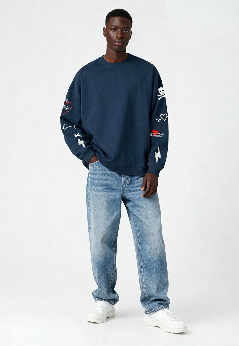 Sweatshirt bleu marine avec des designs brodés sur les manches, featuring des symboles comme des cœurs et des crânes. Associé à un jean bleu clair et des baskets blanches.