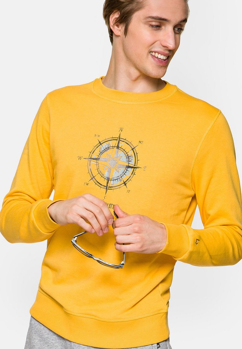 Żółta bluza z miękkiego materiału, z grafiką kompasu, prążkowanymi mankietami i okrągłym dekoltem. Model trzyma okulary przeciwsłoneczne.