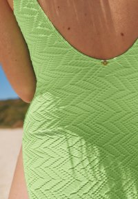 Maillot de bain une pièce texturé vert avec un décolleté dos profond et des motifs en zigzag. Présente un petit accent métallique près du décolleté.