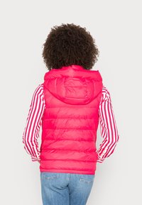 Gilet rouge rembourré avec une capuche, présentant une texture matelassée. Porté par-dessus une chemise à rayures blanches et rouges à manches longues et un jean bleu clair.