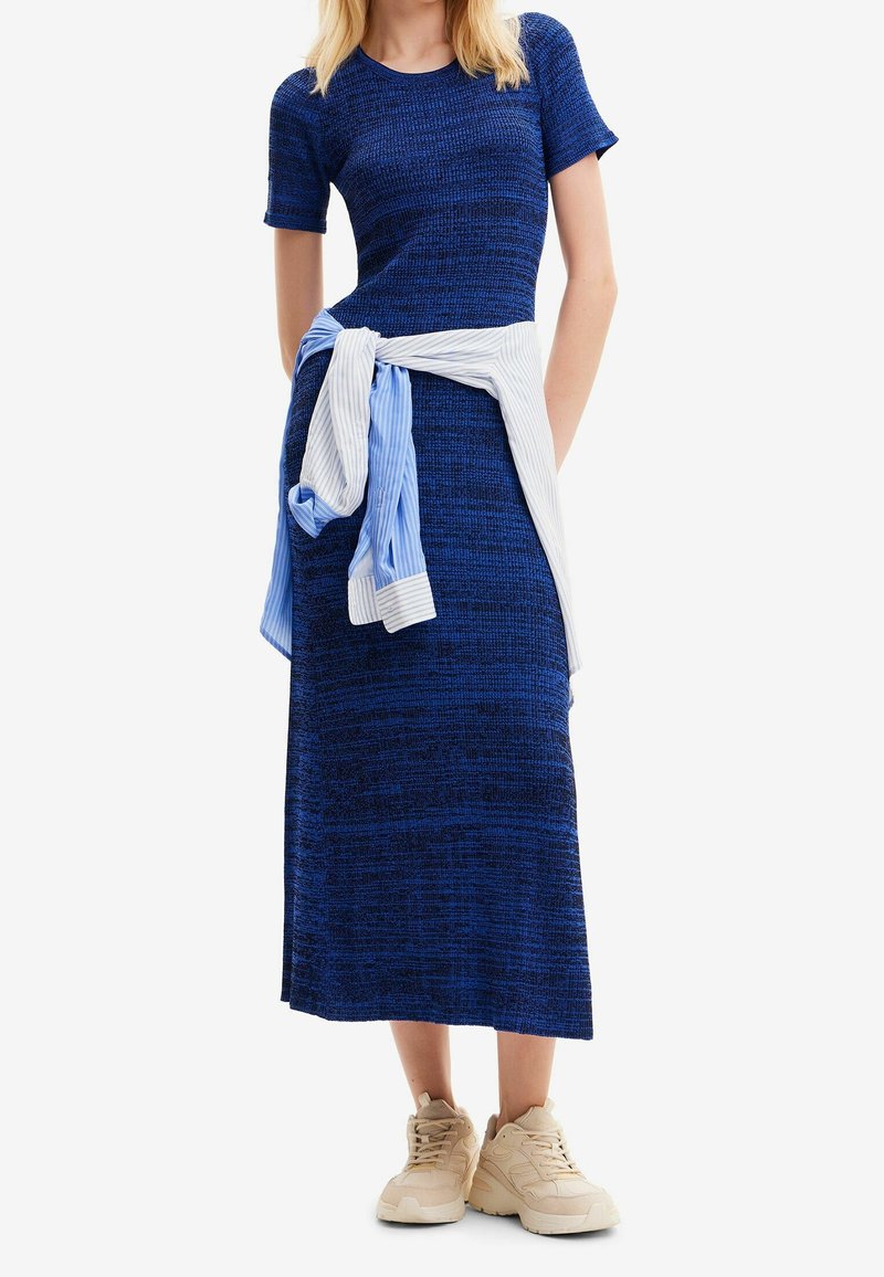 Robe tricotée bleue à manches courtes, texture côtelée et longueur midi, associée à une chemise blanche à rayures bleues nouée à la taille.
