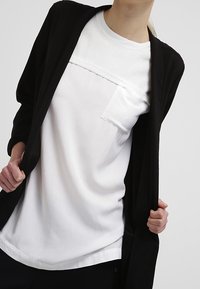 Cardigan aperto nero sovrapposto a una T-shirt bianca con dettaglio testurizzato e taschino. Tessuto morbido con un fit rilassato e scollo rotondo.
