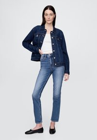 Tamsiai mėlynas džinsinis švarkas su sidabrinėmis detalėmis, derinamas su šviesiai mėlynais džinsais. Modelis dėvi baltą palaidinę ir juodus lygiais pado batais.