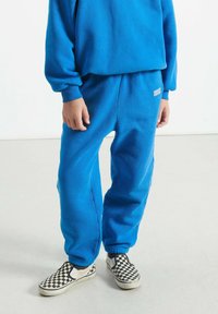 Bērns, kurš valkā zilos sporta bikses un sporta kreklu ar baltām šaha modeļa čības, stāv uz gaišas grīdas.