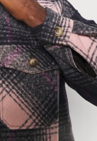 Arm med mørk hudtone iført grå, pink og lilla ternet uldfrakke med brune knapper, hånd delvist inde i lommen på hvid baggrund.