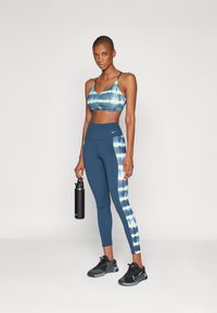 Sutiã desportivo com um padrão em tie-dye azul; leggings de cintura alta em azul marinho sólido, com uma faixa lateral em tie-dye; sapatilhas desportivas pretas.