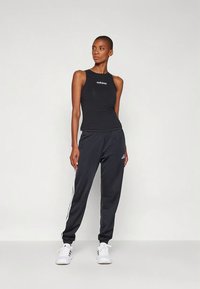 Ženska v črni Adidas brezrokavnik in črnih trenirkah z belimi črtami, ki stoji pred enostavno svetlo ozadje.