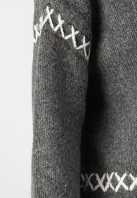 Grauer Wollpullover mit strukturiertem Strickmuster, der weiße Kreuzstichmuster entlang der Oberseite aufweist. Nahtdetails sind an den Ärmeln sichtbar.