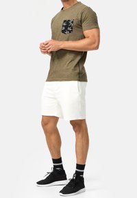 Camiseta de manga corta caqui con un bolsillo negro estampado, combinada con pantalones cortos blancos y zapatillas deportivas y calcetines negros. Diseño sencillo.