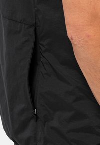Tessuto nero con una texture e dettaglio con zip laterale. Il capospalla ha una vestibilità aderente e presenta una piccola tasca vicino alla cucitura.