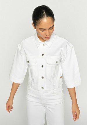 Junge Frau mit dunklen Haaren zu einem glatten Dutt frisiert, trägt eine weiße, geknöpfte, verkürzte Jacke und weiße Hosen, schaut nach unten, die Arme leicht ausgestreckt.