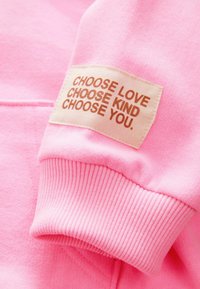 Puño de manga de tela rosa con una etiqueta beige cosida que dice "CHOOSE LOVE CHOOSE KIND CHOOSE YOU" en texto marrón.