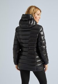 Juodas pūkinis švarkas su blizgiu paviršiumi, su gobtuvu ir horizontaliai siuvinėtu dizainu. Elastingi rankogaliai ir šiek tiek ilgesnė nugaros dalis, kad būtų užtikrintas uždengimas.