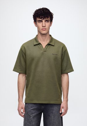 Jeune homme aux cheveux bouclés et foncés portant un polo vert olive à manches courtes et un jean gris foncé, debout sur un fond uni.