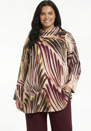 Vrouw glimlachend met een blouse met lange mouwen en hoge hals met abstracte paarse, groene en beige bladpatronen en een donkere broek.